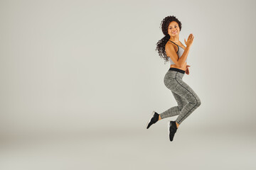 A young African American woman in athletic attire joyfully jumps against a plain gray backdrop in a...