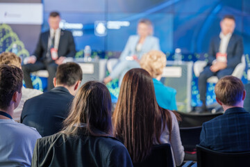 A diverse audience attentively listens to speakers on stage during a business conference. The...