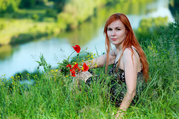 A girl with wild poppies enjoys the view from a hill near the river. Joy, happiness, serenity. Wild poppies by the river.