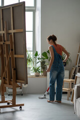 Female artist in denim overall cleaning floor by vacuum cleaner in art workshop in earphones listening music or podcast. Woman make tidy after painting artwork, prepare premises for new project.