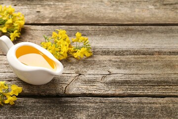 Rapeseed oil in gravy boat and beautiful yellow flowers on wooden table, space for text