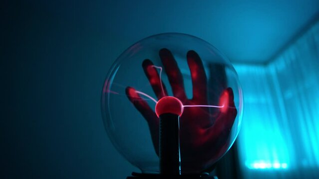 Closeup of evil wizard touching electric plasma ball with luminous flames in dark interior. Low-angle view of Tesla lamp with high voltage lightning, slow motion. Lasers moving from red plasma ball.