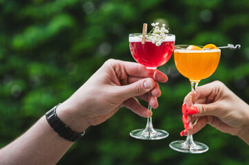Two hands hold stemmed glasses with colorful drinks—one red and one orange—against a blurred green foliage background