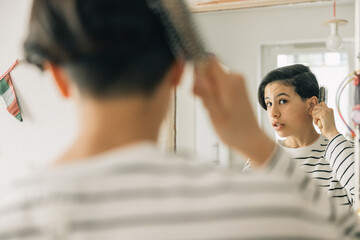 A teenager is brushing her hair in front of a mirror, she is wearing a striped shirt and has short...