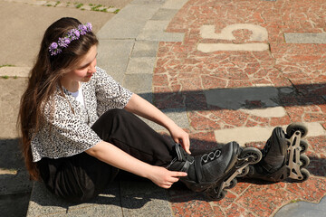 A teenage girl on roller skates with her hair down and a lilac wreath on her head sits on a granite...