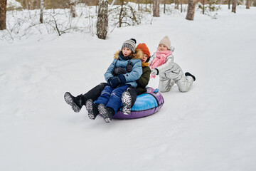 Two happy affectionate children sliding down hill on snow tube while cute girl in winterwear pushing them during play in winter forest