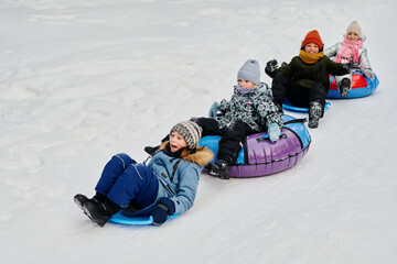 Row of adorable happy children in winter jackets sitting on slides and snow tubes and riding down...