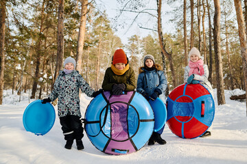 Several happy intercultural children in winterwear holding snow tubes and slides and looking at...