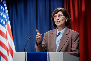 Confident mature female deputy or minister in formal suit standing by platform and pointing...