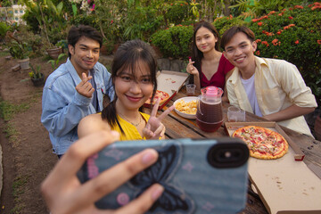 A young asian woman takes a selfie together with three of her friends while enjoying pizza and other snacks during a picnic at the lawn.