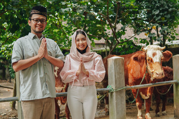 Lovely young Asian Muslim couple doing traditional greeting gesture in front of cows cattle for...