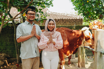 Lovely young Asian Muslim couple doing traditional greeting gesture in front of cows cattle for sacrifices. Eid Al Adha concept.