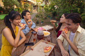 Four young asian friends enjoying pizza and drinks during an outdoor picnic in a lush garden or...