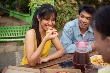 A young woman enjoys eating pizza with her boyfriend and friends during a casual outdoor picnic in...