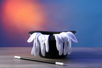 Magician's hat, wand and gloves on wooden table against color background