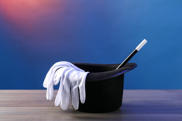Magician's hat, wand and gloves on wooden table against color background