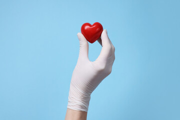 Doctor wearing white medical glove holding decorative heart on light blue background, closeup