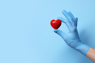 Doctor wearing medical glove holding decorative heart on light blue background, closeup. Space for...
