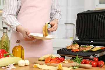 Woman cooking different products with electric grill at wooden table in kitchen, closeup