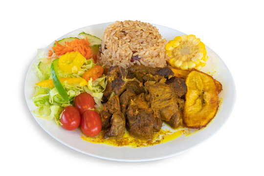 A colorful plate of Caribbean or Jamaican curried goat dinner with rice, corn, plantain and salad isolated on white