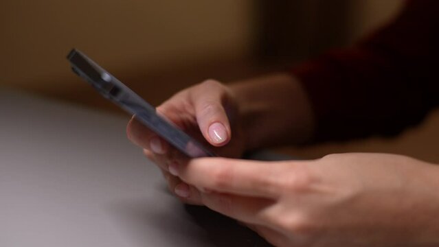 Female hand scrolling browsing on smartphone at night time sitting at table. Close-up of unrecognizable woman using cellphone, browsing social network, scroll through news feed on app on phone..