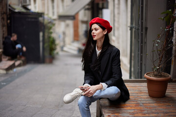 Stylish woman tourist in stylish clothes in a jacket and red beret sitting outside with phone in...