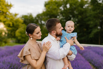 Family with a baby on a lavender field. A woman in brown dress and a man in white skirt with a beard are holding a newborn baby girl in a blue dress in a lavender field. Family, lifestyle concept.