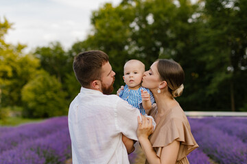 Fototapeta premium Family with a baby on a lavender field. A woman and a man with a beard are holding and kissing a newborn baby girl in a blue dress in a lavender field. Family, lifestyle concept.