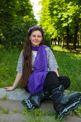 A young girl on roller skates and wearing a purple scarf sits on the grass in a city park in the...