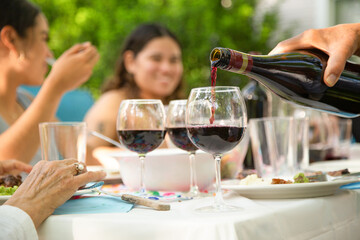 A group of people are sitting at a table with wine glasses and a bottle of wine being poured. Scene is relaxed and social, as the people are enjoying each other's company and sharing a meal together
