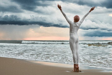 Hairless girl with alopecia in white futuristic suit standing on sea beach stretched out arms to...