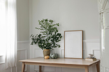 Home office concept. Old books, empty vertical wooden picture frame mockup on office desk, table. Tree branches in vase. Cup of coffee, pumpkins. Working space. Scandinavian interior. Blurred stove