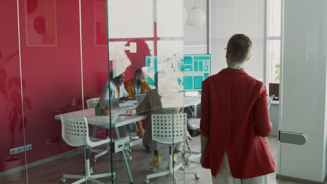 Medium full follow slowmo of Caucasian woman in red jacket walking through glass doors into meeting room at marketing company for project discussion, greeting multiethnic colleagues sitting at table