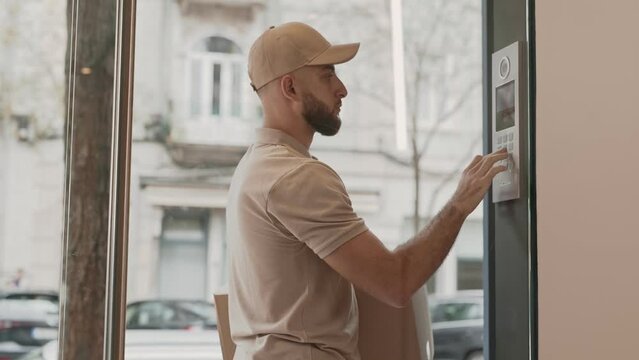 A male delivery courier is calling an apartment from the entrance intercom before entering the building. He holds a parcel and looks attentive, ready to complete the delivery process.