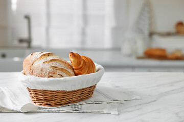 Wicker bread basket with freshly baked loaf and croissant on white marble table in kitchen, space...