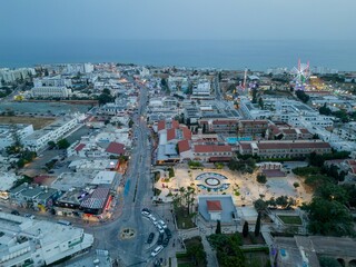 Illuminated Ayia Napa town square with fountain in Cyprus. Aerial view towards high street and the amusement park with colorful lights at dusk.