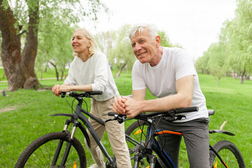 elderly senior couple rides bicycle in the park in the summer and smiles, old gray-haired man and woman are actively resting outdoors, old people practice cycling in forest - Powered by Adobe