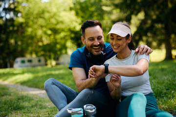 Happy athletic couple using smart watch while relaxing after exercising in nature.