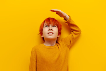 red-haired child boy measures his height and reaches up on a yellow isolated background, a teenager...
