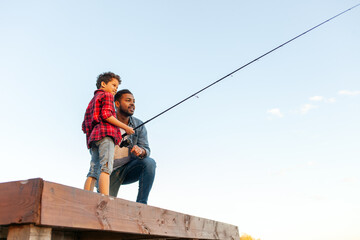 african american father and son fishing in the lake and sitting on wooden pier and holding fishing rod outdoors in summer, man teaches child to fish and relax outdoors on river