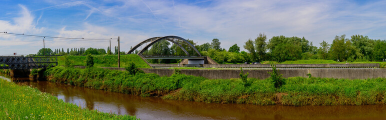 Obraz premium panorama photo of a beautiful landscape with lots of greenery, a river with an arched bridge for transport and a blue sky with clouds.