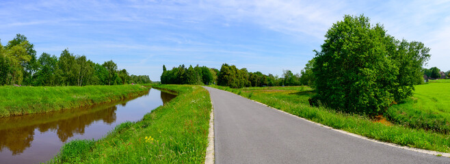 Panoramic photo of a cycle path next to a calm river surrounded by green forests and a clear blue...