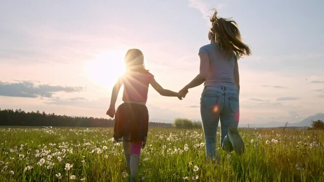 Slow motion shot of a mother and her little daughter running holding hands across the beautiful green grassland at sunset, rear view.
