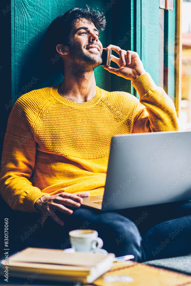 Poster happy handsome young man having friendly mobile conversation on smartphone while working at laptop c