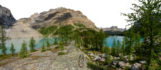 A large rocky mountain with two light blue lakes in the midground and rocks and trees in the foreground. The sky is transparent.
