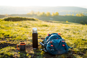 A backpack, thermos, and mug rest on a grassy hill within a natural landscape. The highland scenery...
