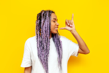 cheerful african american woman with colored dreadlocks showing gesture of smoking marijuana and hemp on yellow isolated background, hipster girl with purple braids and unique hairstyle showing ganja