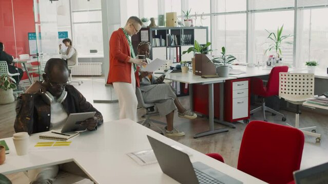 Full dolly-out shot of diverse male and female advertising agency employees working in bright spacious open plan office - discussing report, preparing presentation, using digital tablet, headphones