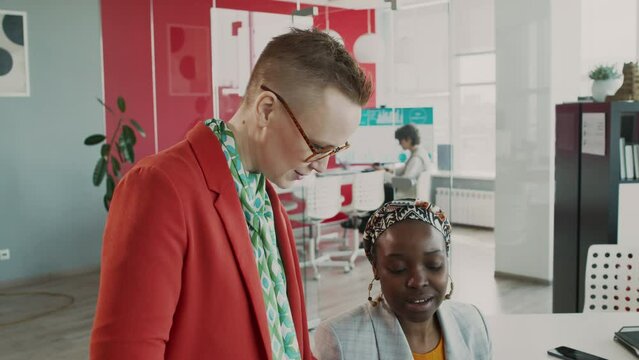 Medium tilt shot of young Caucasian female marketing manager in red jacket, stylish blouse discussing marketing report with African American specialist in ethnic turban in corporate office