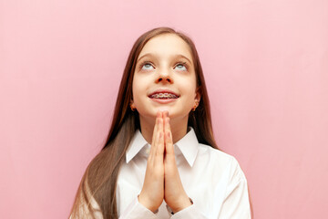 cheerful teenage girl with braces asks and prays on pink isolated background, child pleads and thanks and shows gesture of faith and hope